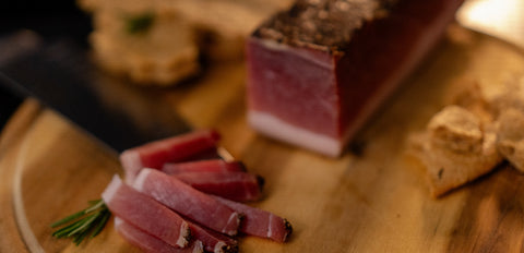 Angeschnittener Speck auf rustikalem Holzbrett mit dünnen Scheiben, Rosmarin und Brot