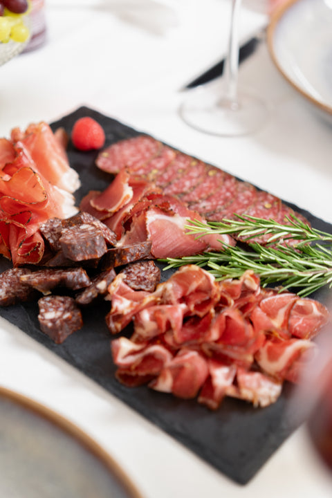 Aufgeschnittener Schinken und Salami dekorativ auf einer Schieferplatte mit Rosmarin und einer Himbeere